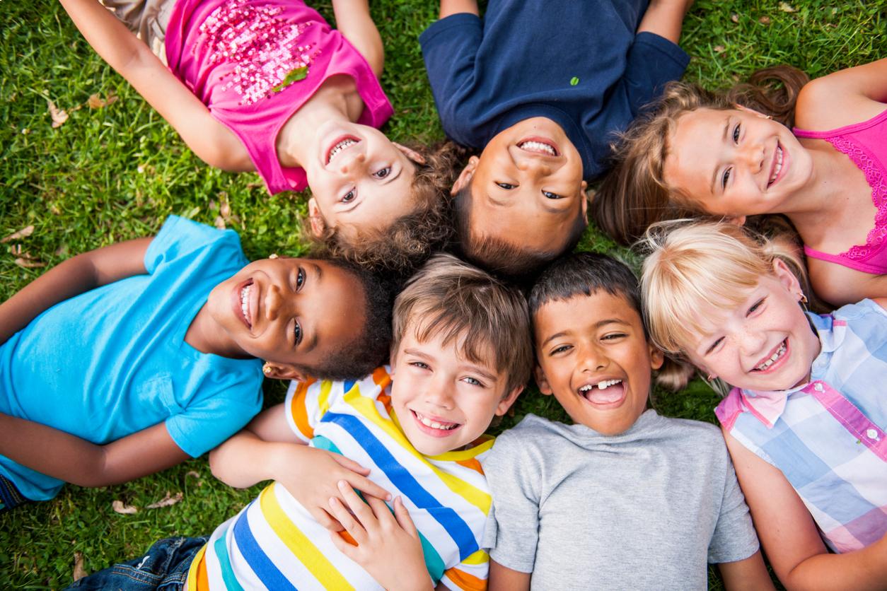 Group of children lying on grass in circle, smiling at camera, outdoor setting.