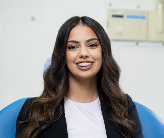 Woman smiling at camera in dental office wearing blue jacket and white shirt.