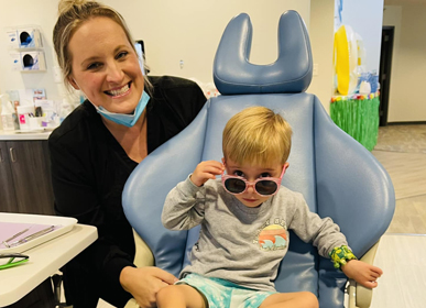 Female dentist with young child in dental chair wearing sunglasses at pediatric dental office.