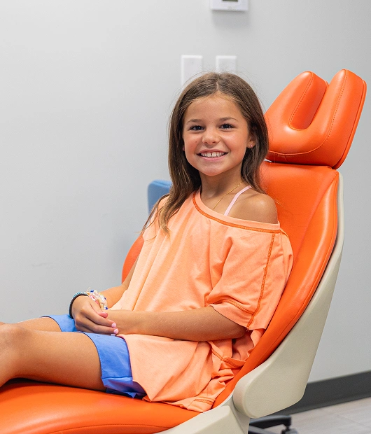 Young girl sitting in orange dental chair at pediatric dental office, smiling at camera.