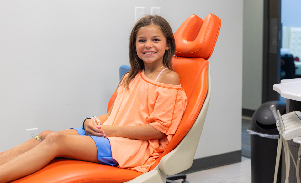 Young girl sitting in orange dental chair, smiling at pediatric dental office.