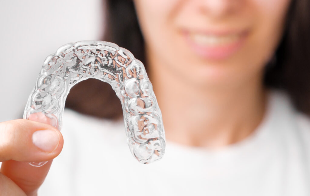 A person holds a clear dental aligner in focus. The background shows a subtle smile, suggesting satisfaction and progress in dental care.