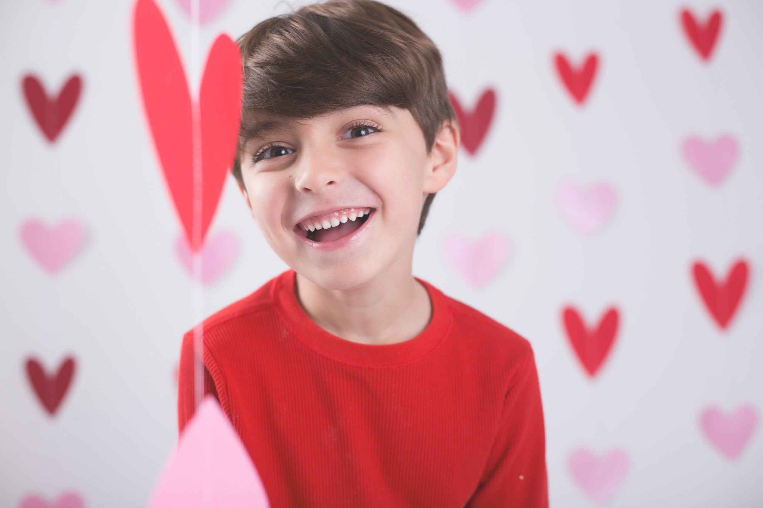 Smiling child in a red shirt surrounded by red and pink heart decorations on a white background, conveying joy and warmth.