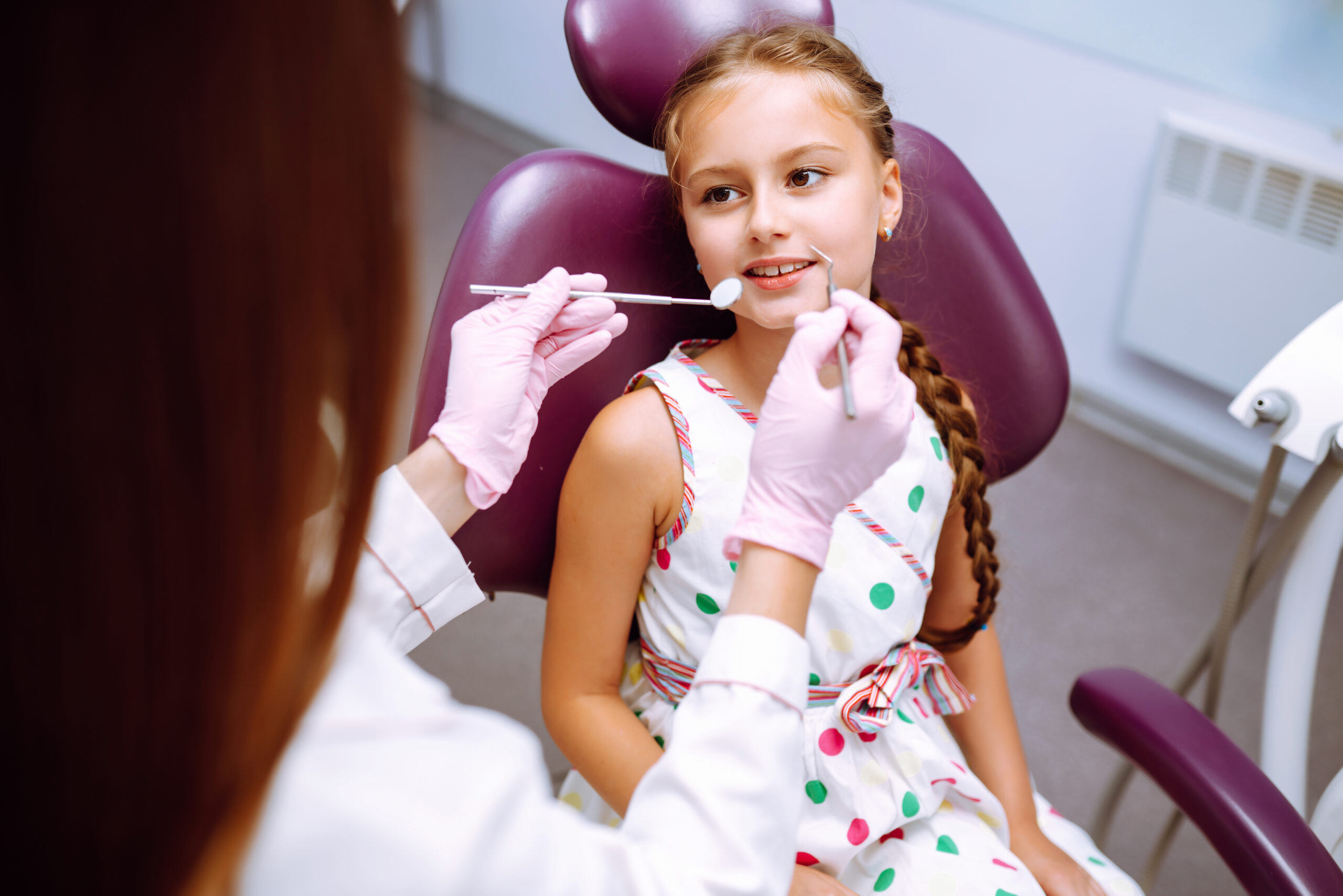 A young girl in a polka dot dress, smiling in a dentist's chair while the dentist, in pink gloves, examines her teeth with a dental mirror.